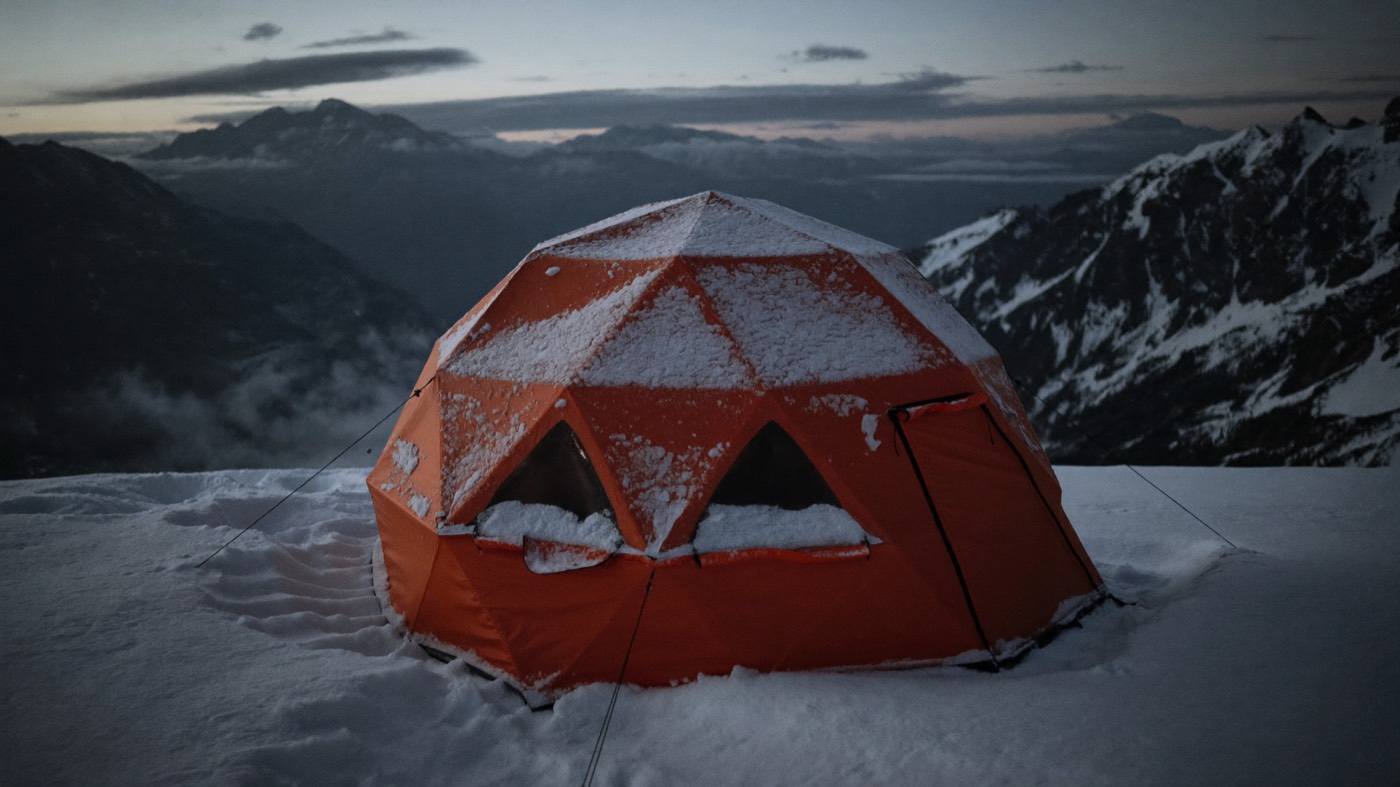 A geodesic dome tent braced on a wind-scoured alpine col under a bank of grey cloud.
