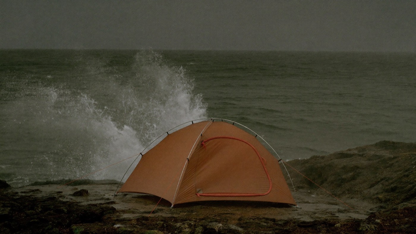 A low aerodynamic tent staked into a rain-swept coastal headland, sea spray blowing inland.