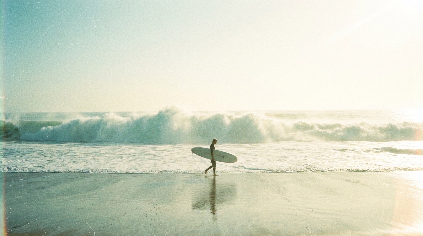 The sun-bleached hero photograph: a surfer carrying a longboard along wet sand at noon.